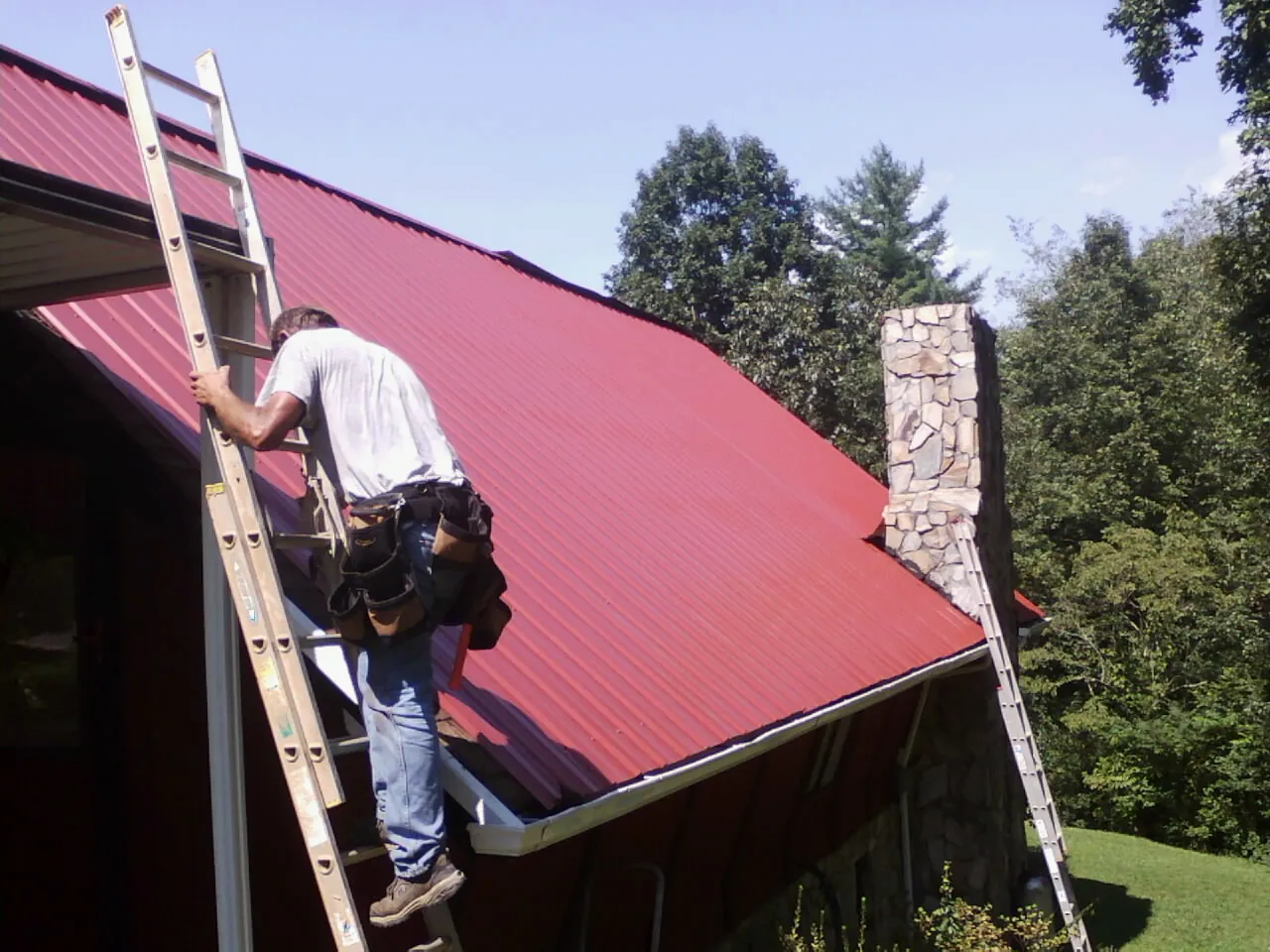 Roofer on a ladder beside a red metal roof and stone chimney — Soffit & Fascia Repair by Roofing Brothers in Moosup